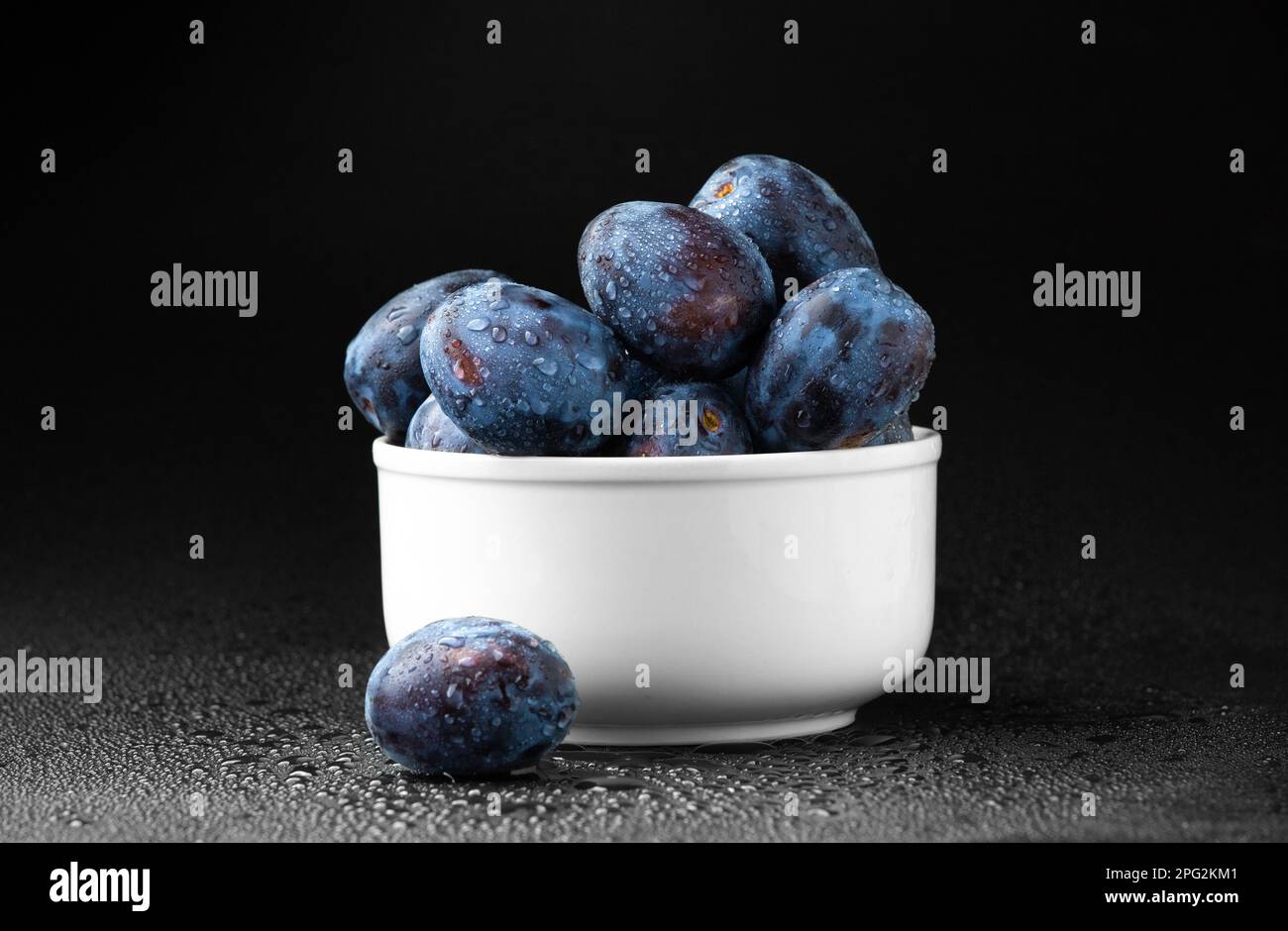 Prune bleue dans une assiette blanche avec gouttes d'eau sur fond sombre. Fruits diététiques utiles. Banque D'Images