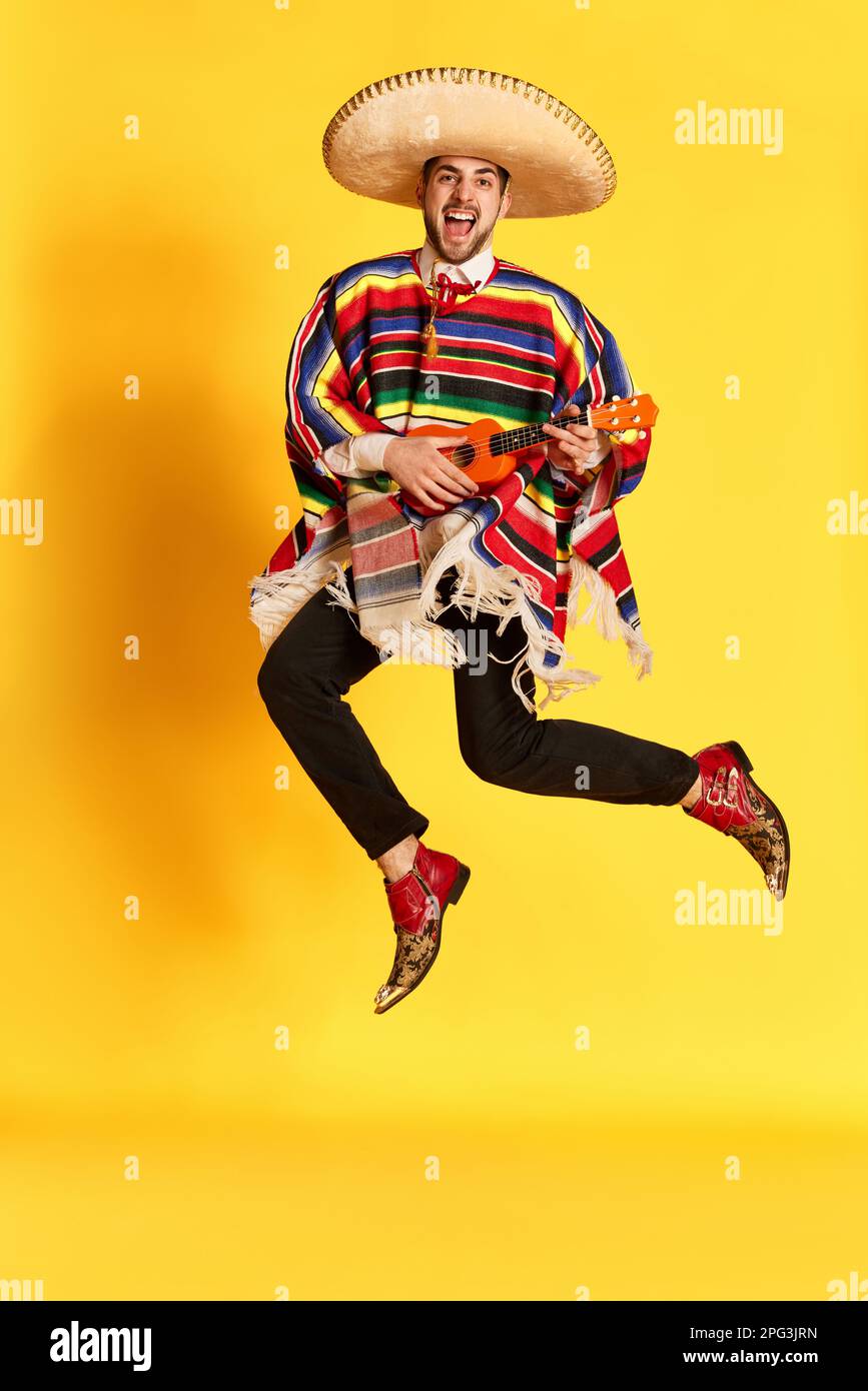 Jeune homme émotif en poncho coloré et sombrero posant, jouant de la guitare et sautant sur fond jaune studio Banque D'Images