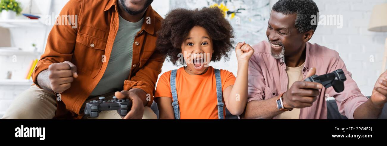 KIEV, UKRAINE - 17 JUILLET 2021: Excitée enfant afro-américain montrant oui geste près des parents avec des joysticks à la maison, bannière Banque D'Images