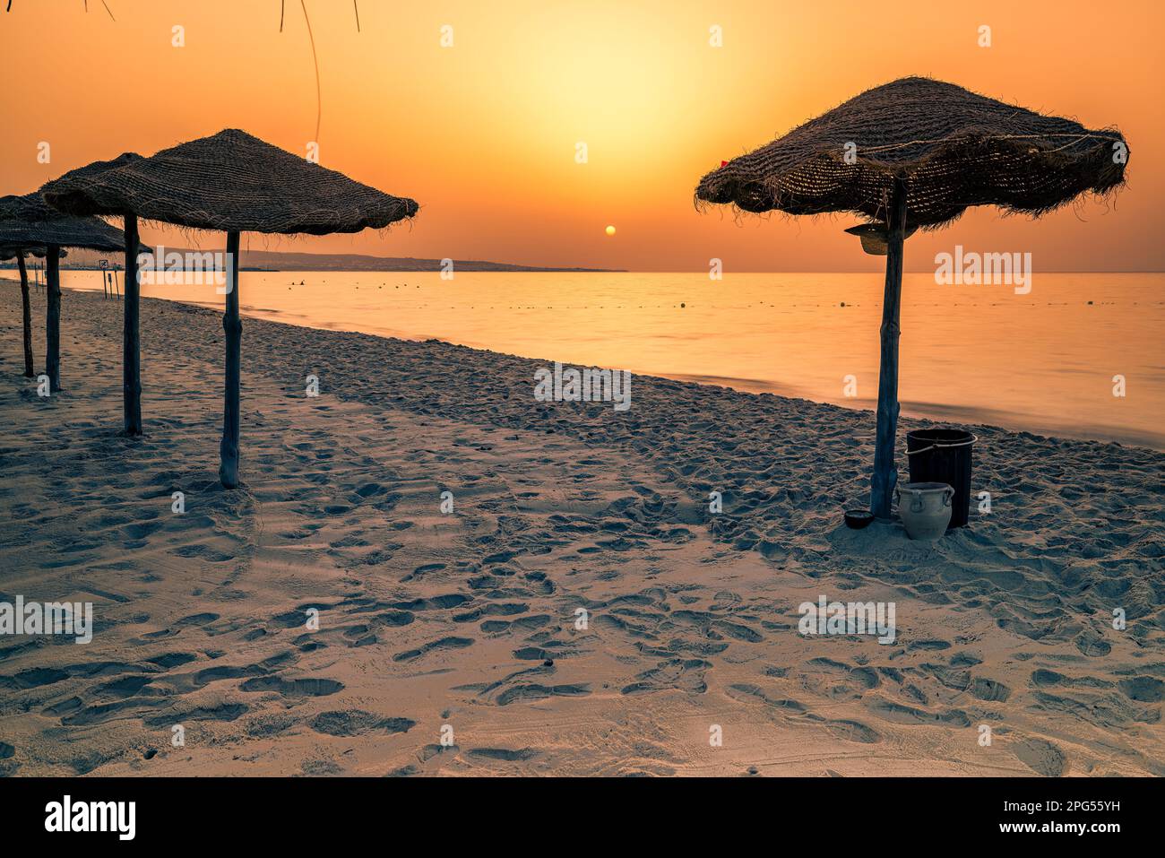 Découvrez la beauté sereine d'une plage tunisienne au coucher du soleil, avec de douces vagues qui s'écrasent contre la rive et deux parasols en bois debout sous un Banque D'Images