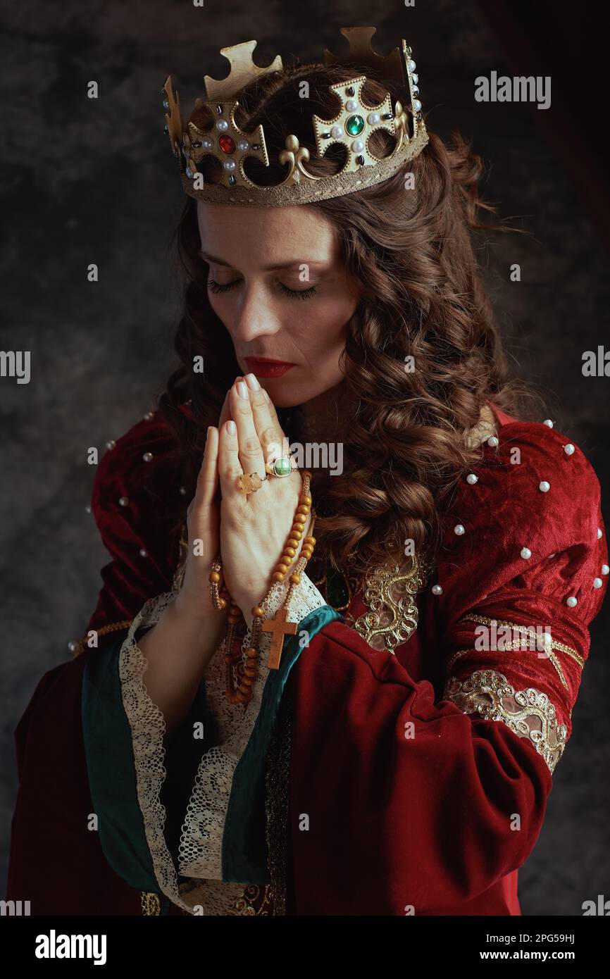 reine médiévale en robe rouge avec rosaire et couronne priant sur fond gris foncé. Banque D'Images