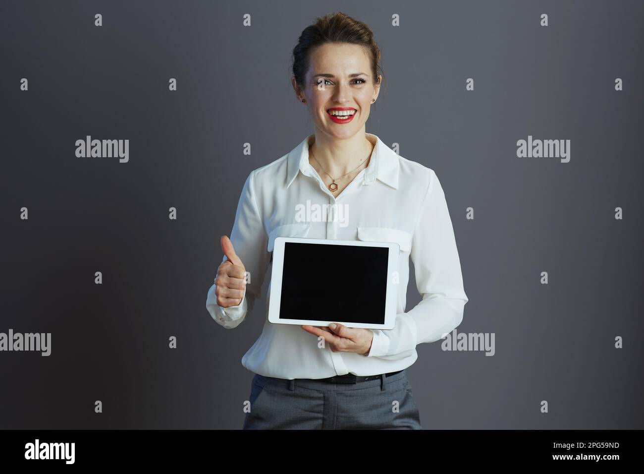 Souriante élégante petite entreprise propriétaire femme dans un chemisier blanc montrant tablette PC écran vide et pouces vers le haut sur fond gris. Banque D'Images