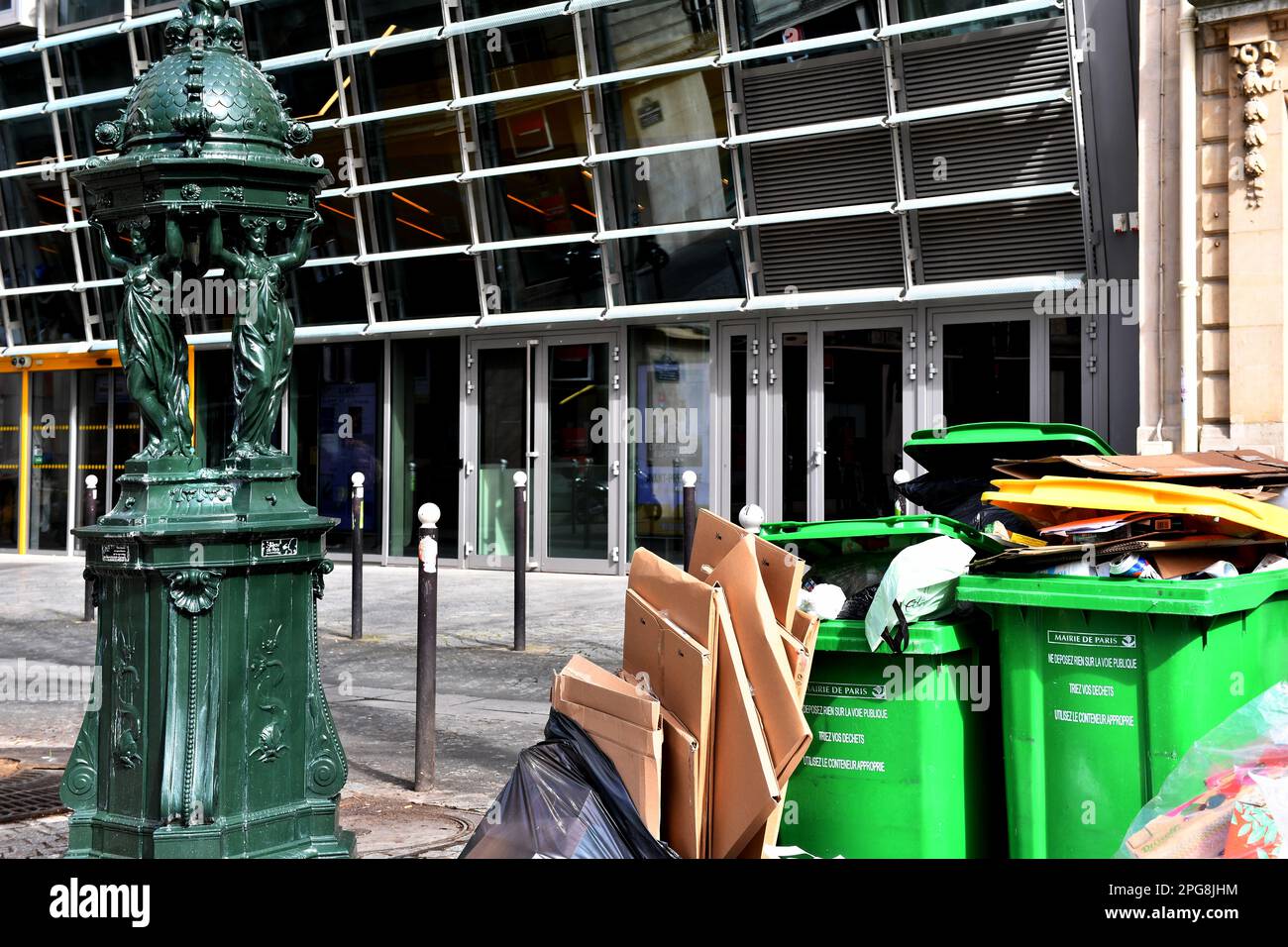 Une fontaine Wallace verte classique à côté de bacs à roulettes verts et de cartons empilés sur un trottoir à Paris, France Banque D'Images