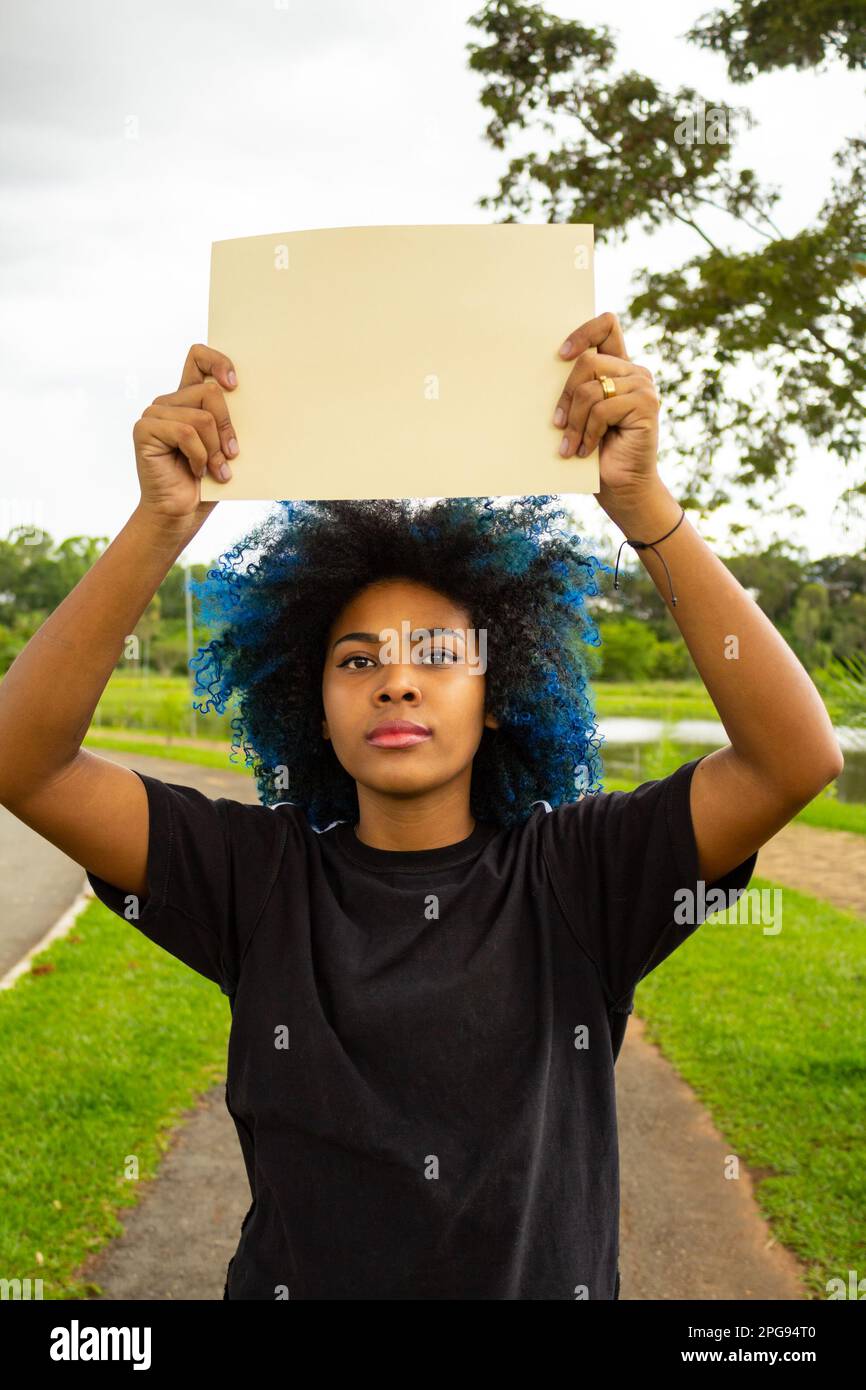 Goiania, Goias, Brésil – 21 mars 2023: Une jeune femme, avec des cheveux bleus teints, tenant un signe blanc, avec un paysage en arrière-plan. Banque D'Images