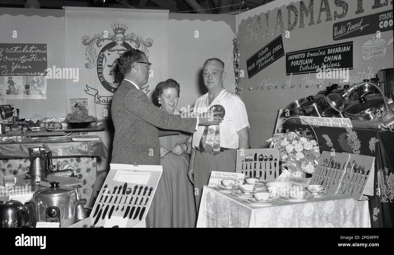 1956, historique, sur un stand d'exposition pour les distributeurs de batterie de cuisine Saladmaster, un couple debout, tandis que l'homme est présenté avec une rosette célébrant le 50th anniversaire de la foire du comté de Lane, Eugene; Oregon, États-Unis. Des couverts en acier inoxydable et de la porcelaine bavaroise sont exposés. L'entreprise a commencé en 1947 avec un seul produit - la machine Saladmaster - et a élargi sa gamme de récipients pour devenir l'une des 100 premières entreprises du Texas à l'horizon 1970s. Banque D'Images