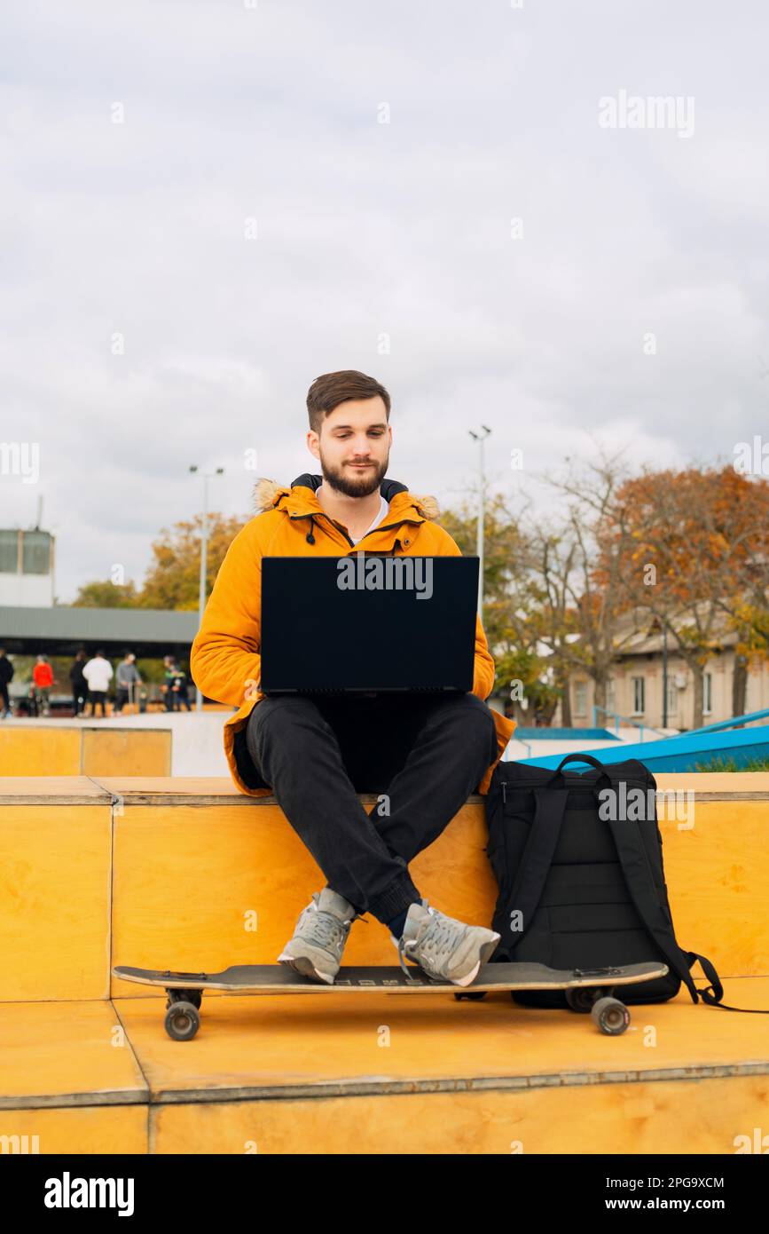 Élégant barbu décontracté jeune homme indépendant travaillant avec un ordinateur portable moderne et utilisant dans un smartphone tout en étant assis sur un banc avec une longue planche à l'extérieur Banque D'Images