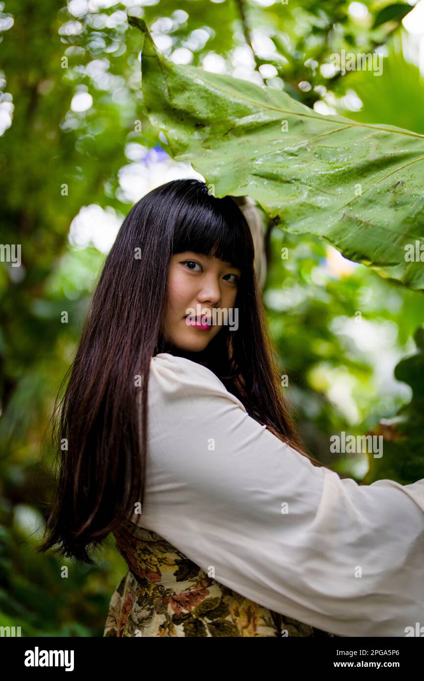 Belle jeune femme asiatique avec des Bangs et des cheveux longs tenant une grande feuille au-dessus d'elle dans une serre | Conservatoire de fleurs Banque D'Images