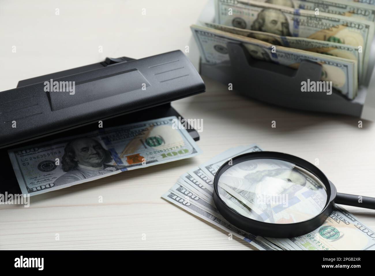Détecteur de devises moderne avec billets de banque en dollars et loupe sur table blanche en bois. Appareil d'examen de l'argent Banque D'Images