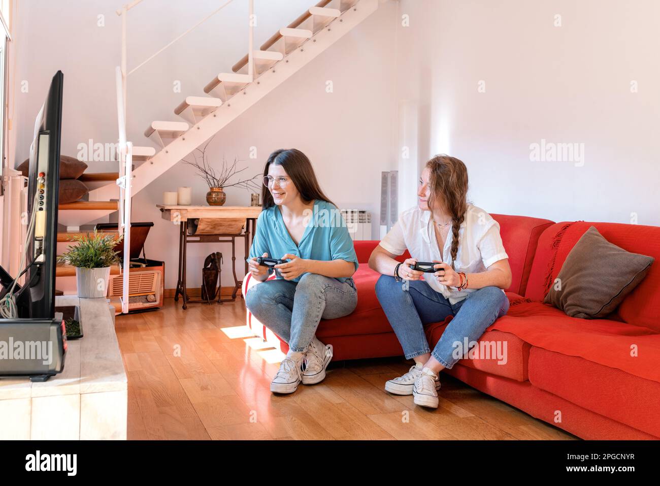 Des jeunes amies joyeuses se détendent sur un confortable canapé rouge avec manettes de jeu vidéo en mains et regardant l'écran de la console de télévision pendant que pla Banque D'Images