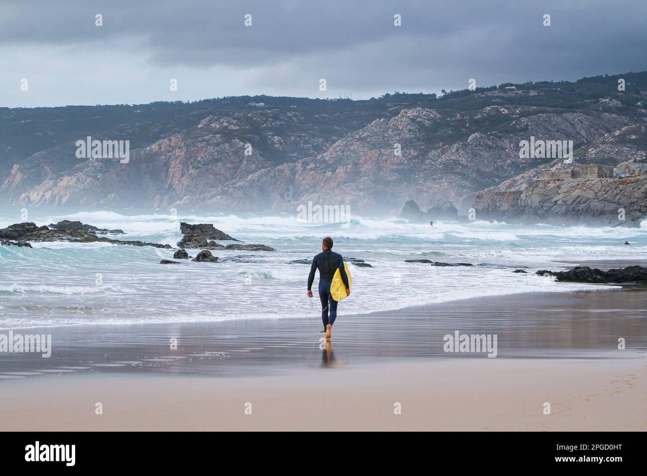 Un surfeur mâle marchant sur une plage de sable vers l'océan transportant une planche de surf sous son bras. Banque D'Images