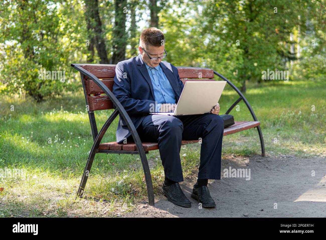 Beau homme d'affaires en costume bleu assis sur le banc dans le parc d'été travaillant à distance sur ordinateur portable. l'homme d'affaires se repose avec un ordinateur portable dans le parc Banque D'Images