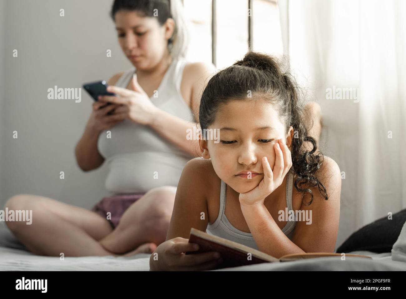 petite fille latine, lisant un livre avec une grande concentration, allongé sur son lit tandis que sa mère regarde son téléphone cellulaire. Banque D'Images