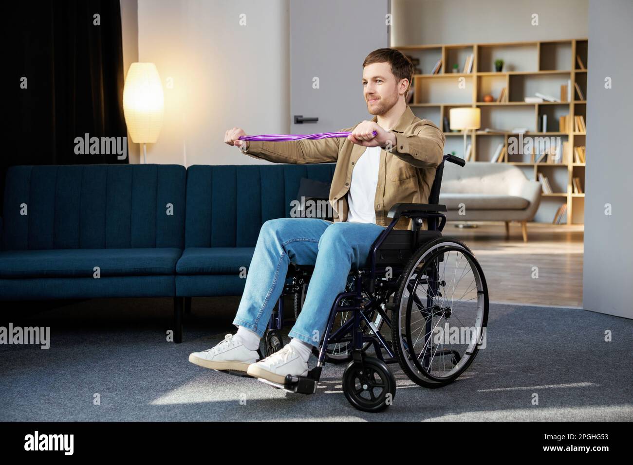 Jeune homme handicapé en fauteuil roulant faisant des exercices avec un élastique à la maison. Un homme handicapé de race blanche s'entraîner dans la salle de séjour. Banque D'Images