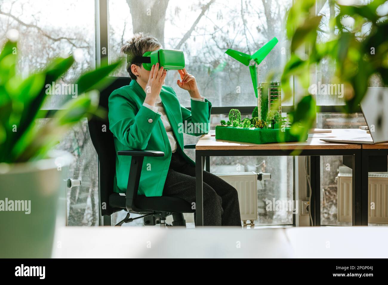 Femme d'affaires portant des lunettes VR assis avec une éolienne et des modèles architecturaux biophiles au bureau Banque D'Images