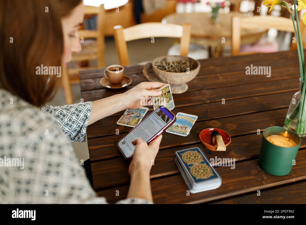 Belle jeune femme devine sur des cartes avec tarot, runes sur table en bois et utilise une application en ligne dans le téléphone pour interpréter la divination, intérieur de maison, bougies et herbes sèches. Banque D'Images