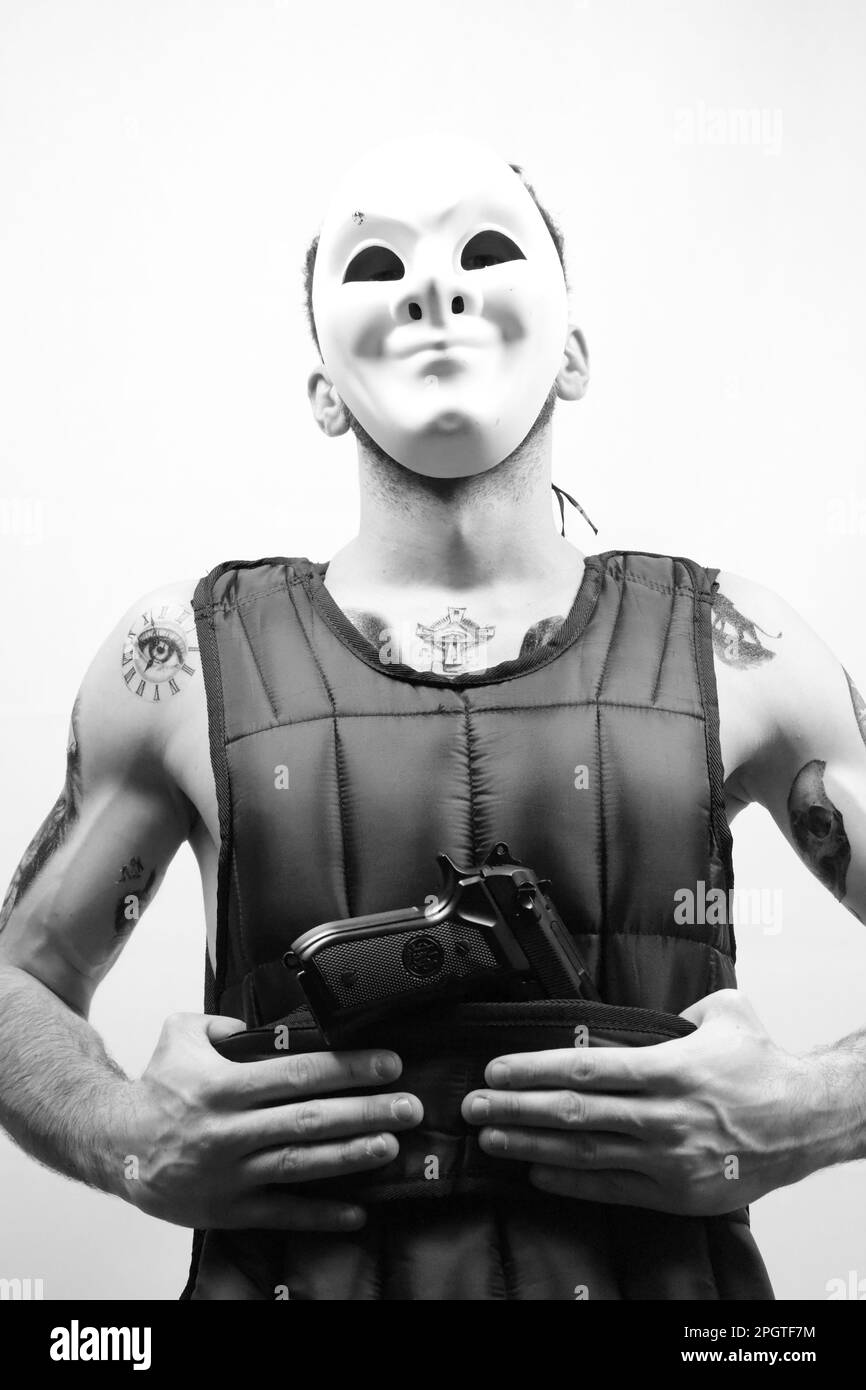 Échelle de gris d'un homme portant un masque facial, avec de nombreux tatouages visibles, tenant un pistolet dans un geste menaçant Banque D'Images