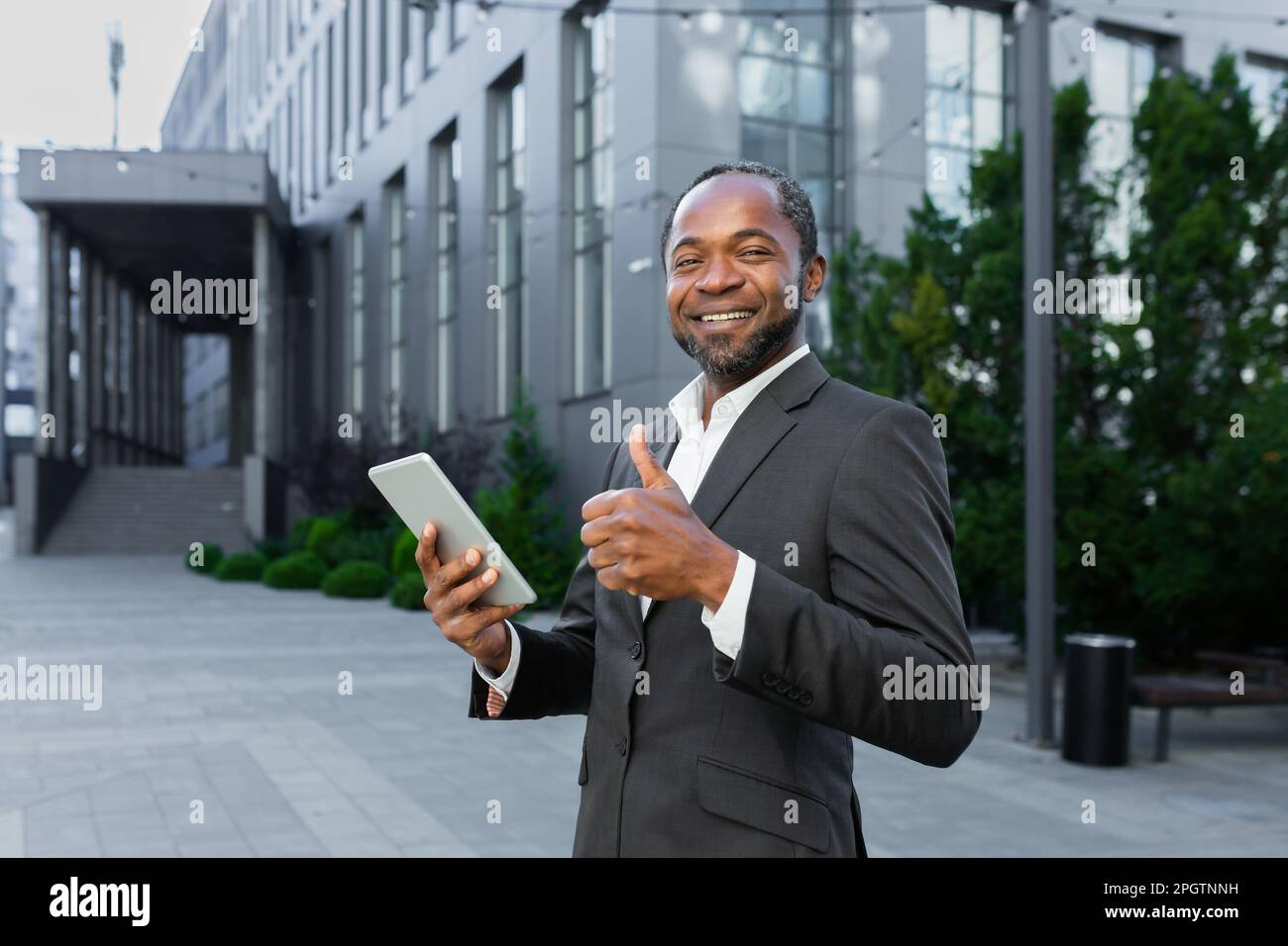 Portrait d'un homme d'affaires noir souriant et réussi debout près d'un bureau et travaillant sur une tablette. Regarde l'appareil photo et pointe un super doigt. Banque D'Images