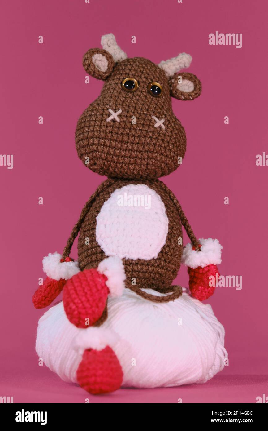 Amigurumi un taureau brun porte des moufles et des chaussons rouges et est assis sur une boule de fil blanche sur fond rose. Jouet bricolage doux en coton naturel et W Banque D'Images