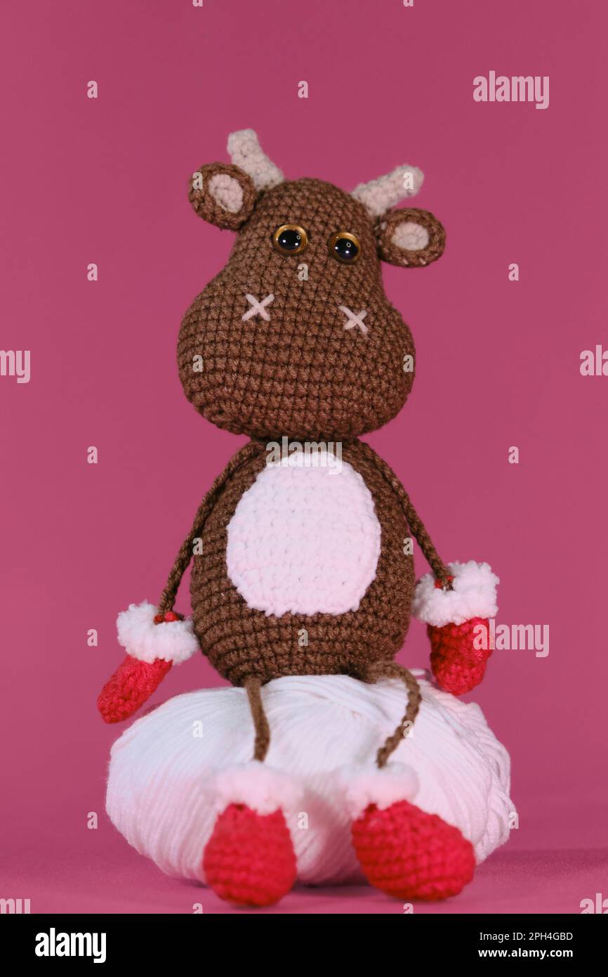 Amigurumi un taureau brun porte des moufles et des chaussons rouges et est assis sur une boule de fil blanche sur fond rose. Jouet bricolage doux en coton naturel et W Banque D'Images