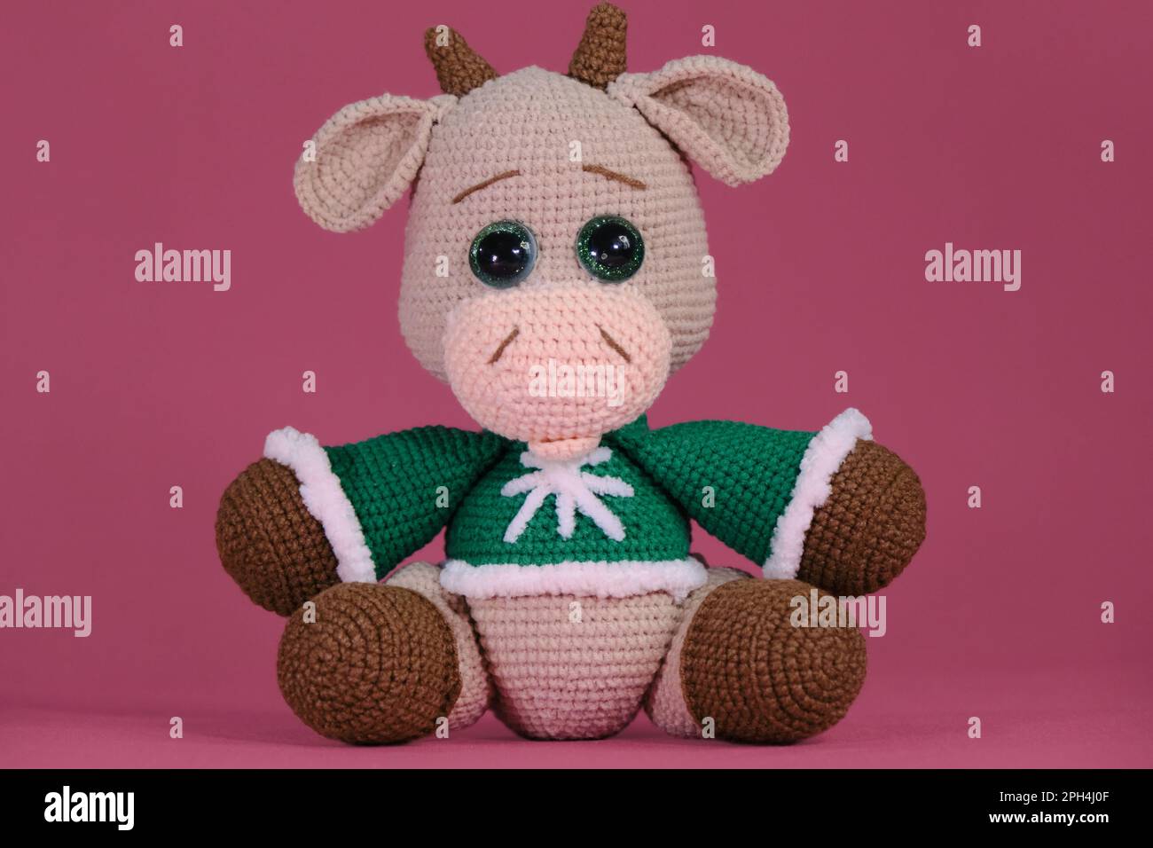 Jolie petite vache à crochettée, art fait à la main Amigurumi un taureau brun porte un chandail vert et est assis sur fond rose. Un jouet doux fait de bricolage naturel Banque D'Images