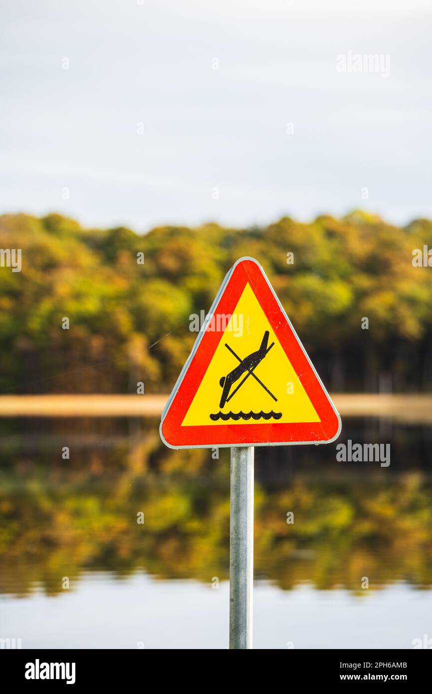 Un panneau d'avertissement jaune vif en forme de triangle interdisant la plongée dans un lac suédois paisible, soulignant l'importance de la communication Banque D'Images