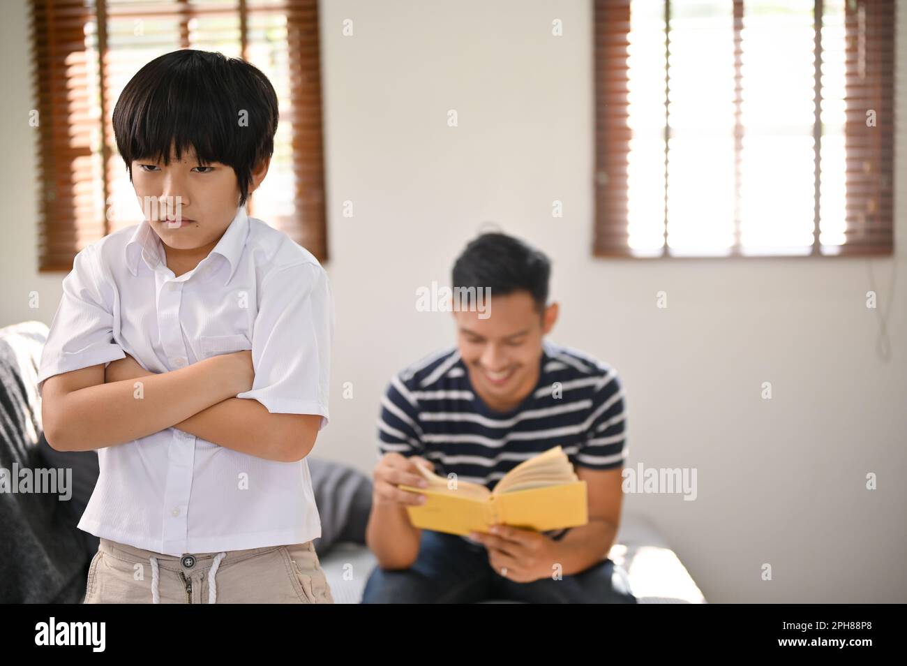 Un jeune asiatique en colère et contrarié se tient avec ses bras croisés dans le salon, se sentant seul et ignoré par son père, qui est en train de lire un livre Banque D'Images