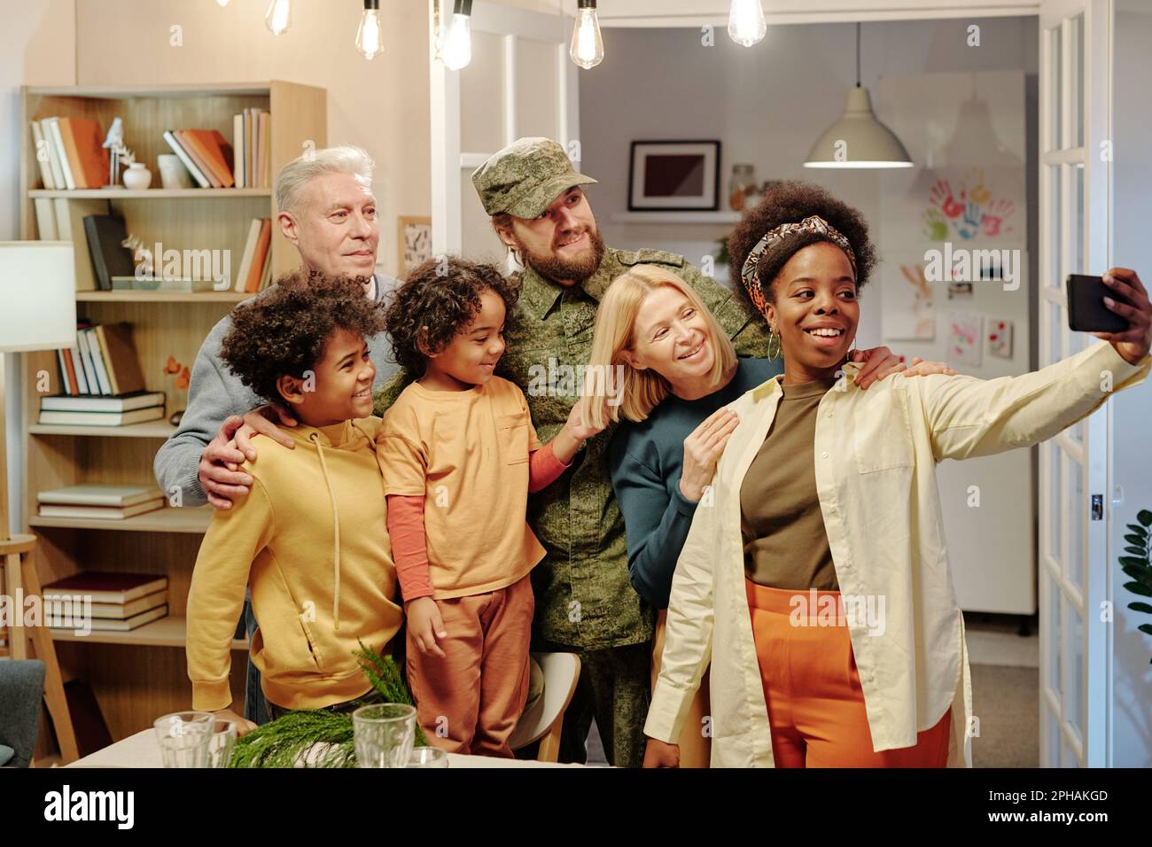 Bonne famille interculturelle de six personnes prenant selfie devant l'appareil photo tout en se tenant dans le salon contre des étagères avec des livres et une porte ouverte Banque D'Images