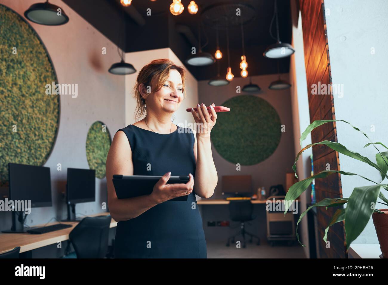 Femme d'affaires ayant un appel professionnel travaillant sur une tablette au bureau. Femme mûre et occupée utilisant un ordinateur tactile debout près de la fenêtre dans un intérieur moderne. FEMA Banque D'Images