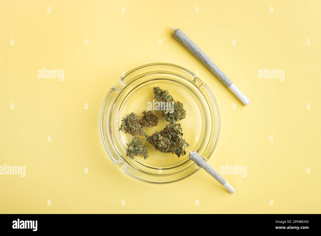 Les bourgeons secs de marijuana médicale se trouvent dans un cendrier en verre, à côté de deux articulations de cannabis de taille différente, sur un fond jaune vif. Beaucoup d'espace vide Banque D'Images