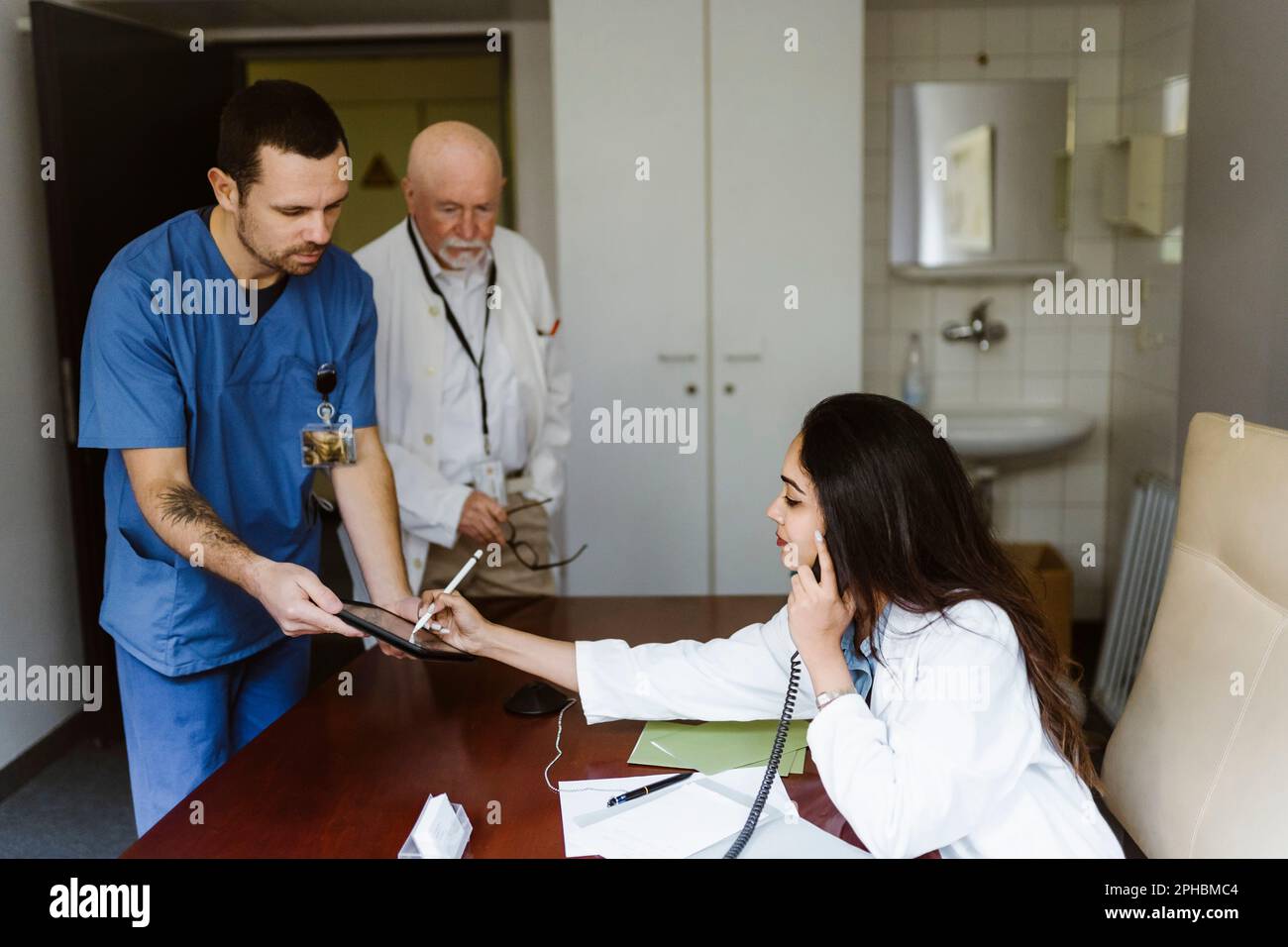Femme médecin parlant par téléphone tout en signant sur une tablette numérique tenue par une infirmière dans le bureau de l'hôpital Banque D'Images