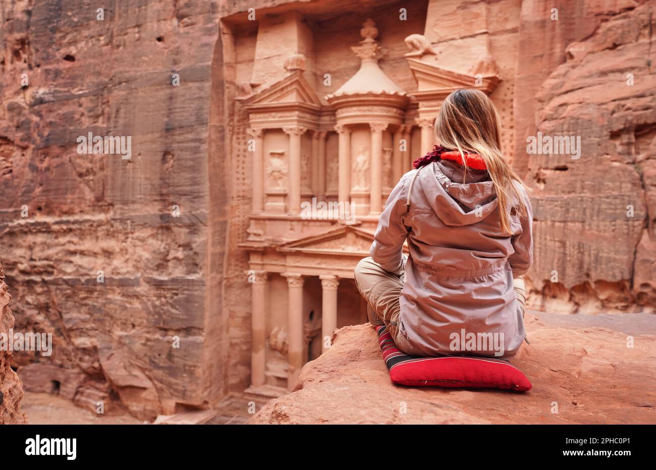 Jeune femme en veste d'hiver est assise sur un petit oreiller rouge au point de vue en regardant Al-Khazneh - treausry, attraction principale de Petra, Jordanie. Vue de behin Banque D'Images