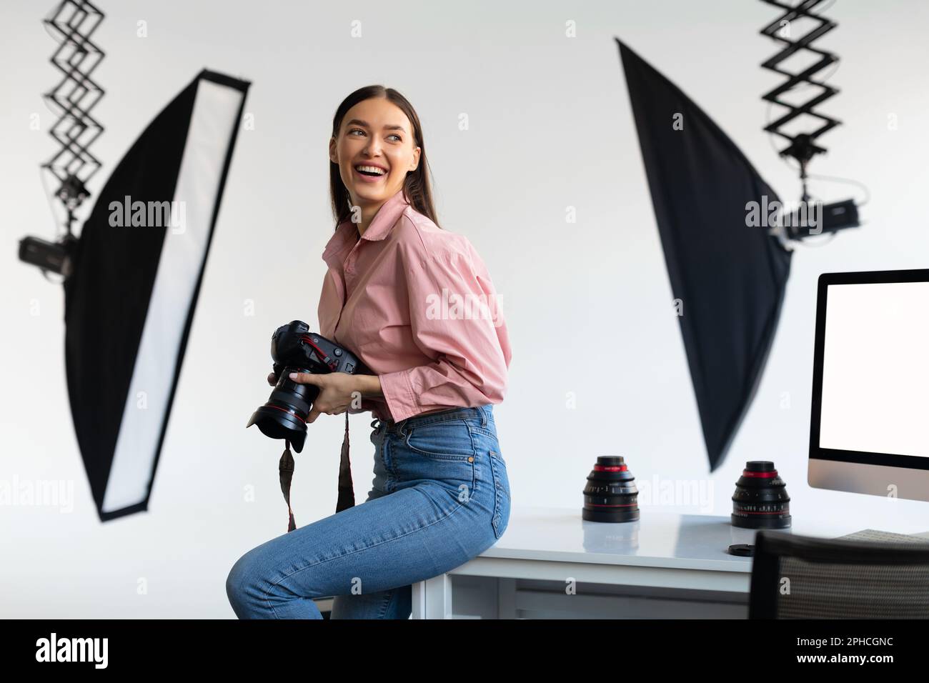 Une photographe excitée assise sur une table, tenant son appareil photo reflex numérique et souriant, à l'intérieur de photostudio Banque D'Images