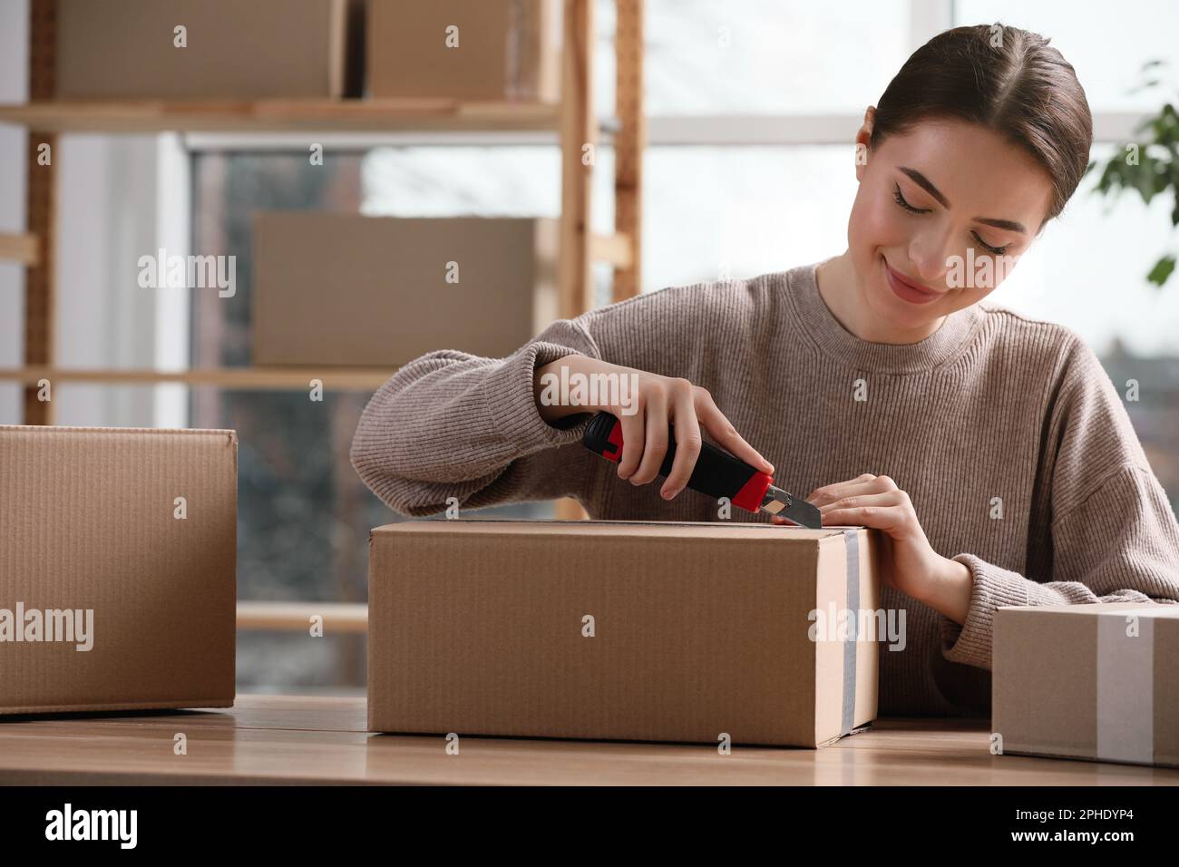 Jeune femme utilisant un couteau utilitaire pour ouvrir la parcelle sur une table en bois à l'intérieur Banque D'Images