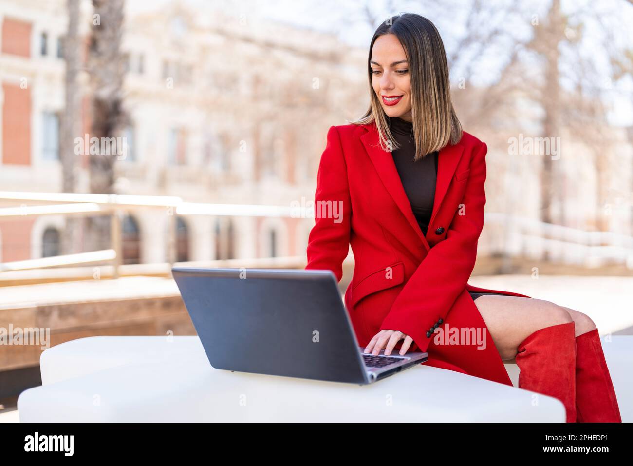 Bonne jeune femme dans les cheveux blond foncé et rouge à lèvres assis à la table et regardant l'écran d'ordinateur portable sur un arrière-plan flou Banque D'Images