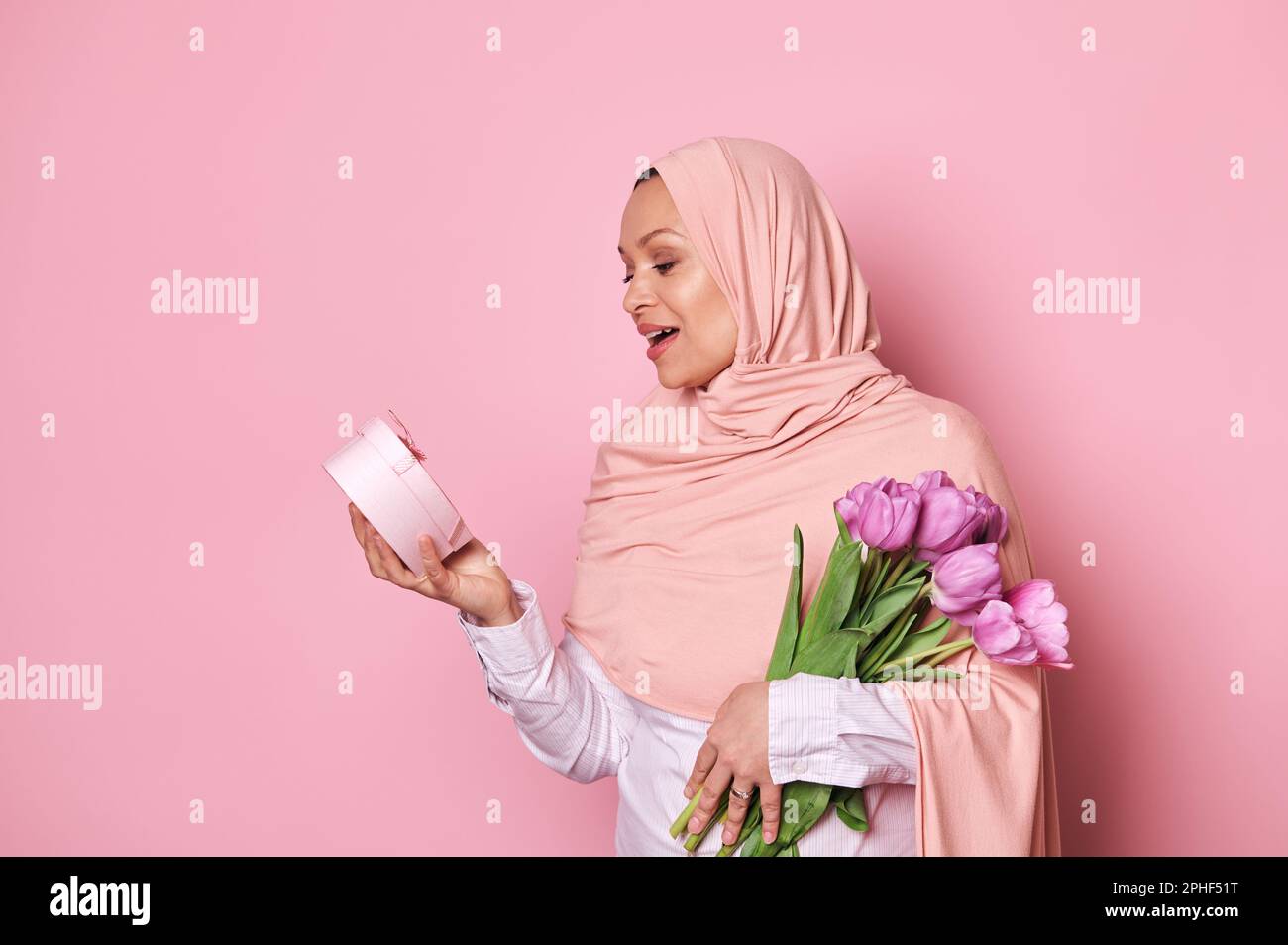 Surprise musulmane jolie femme en hijab rose, tenant un bouquet de tulipes, exprimant l'étonnement tout en obtenant un cadeau heureux dans une boîte rose pour fe Banque D'Images