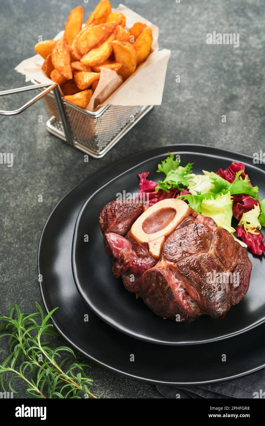 Steaks Osso Buco. Bœuf grillé Osso Buco ou steak de viande de portreuse avec garniture de salade et de frites sur un panneau de marbre noir sur du vieux bois Banque D'Images