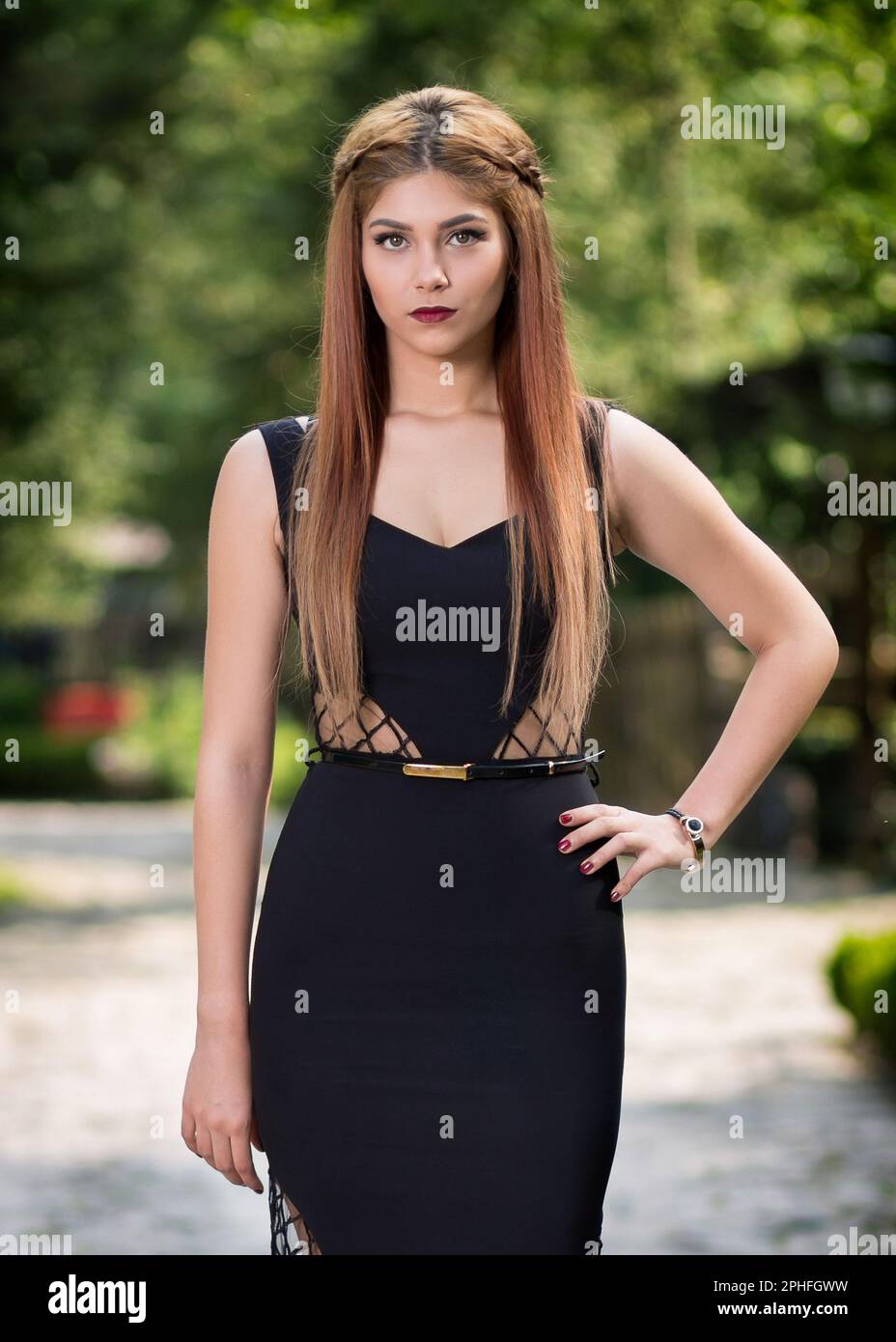 Une jeune femme aux longs cheveux de brunette, portant une robe de soirée noire, se dresse sur une rue pavée dans une posture gracieuse Banque D'Images