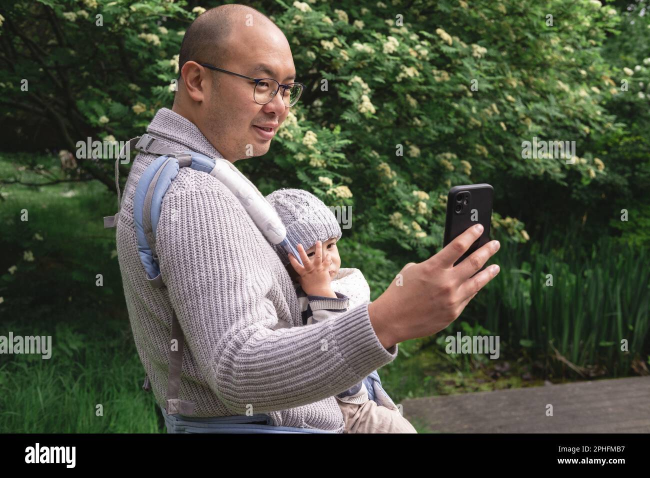 Un nouveau père souriant portant son bébé avec un porte-bébé prenant une photo de selfie avec son téléphone cellulaire dans la nature au parc au printemps. Briti Banque D'Images