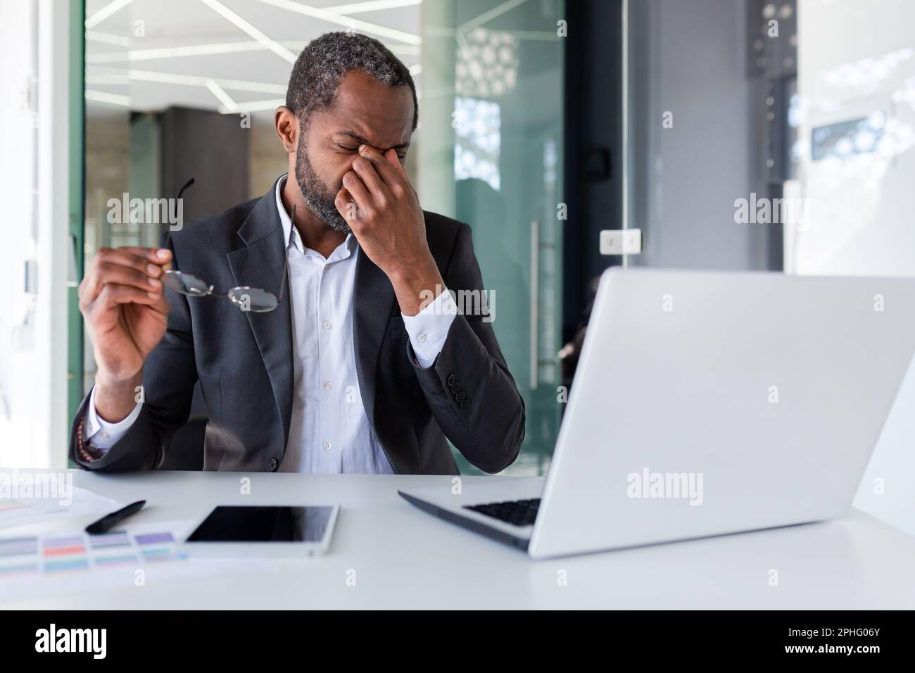 Un homme surtravaillé sur le lieu de travail à l'intérieur du bureau, un homme d'affaires afro-américain travaillant assis avec un ordinateur portable au travail, un patron qui délasse des lunettes et massante les yeux. Banque D'Images