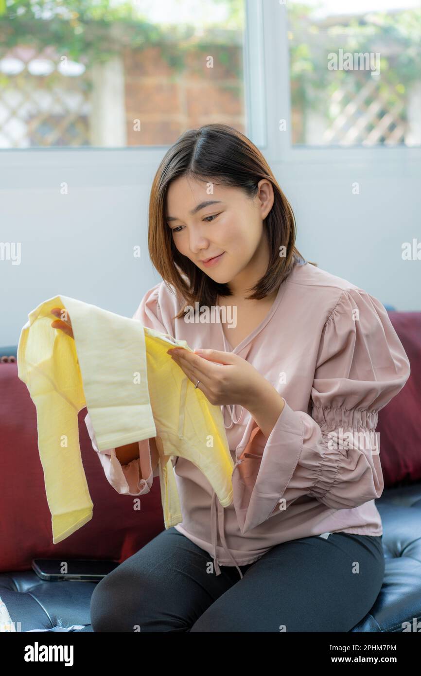 Une jeune femme asiatique heureuse, vêtue de vêtements de maternité, souriant et regardant un nouveau vêtement de bébé qu'elle tient tout en étant assise dans sa maison lumineuse Banque D'Images