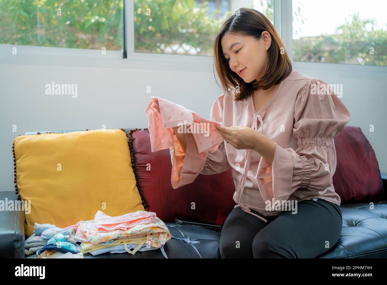 Une jeune femme asiatique heureuse, vêtue de vêtements de maternité, souriant et regardant un nouveau vêtement de bébé qu'elle tient tout en étant assise dans sa maison lumineuse Banque D'Images
