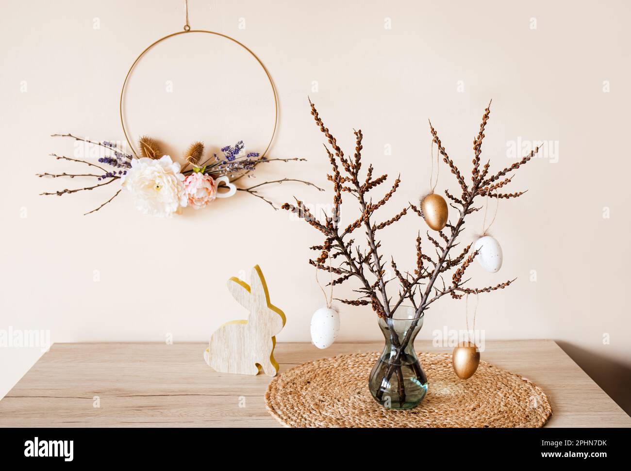 Décoration de Pâques maison. Guirlande de fleurs rondes à motif ressort fait main accrochée au mur, branches d'arbre sur vase avec œufs suspendus. Belle tonalité beige chaud terrestre Banque D'Images