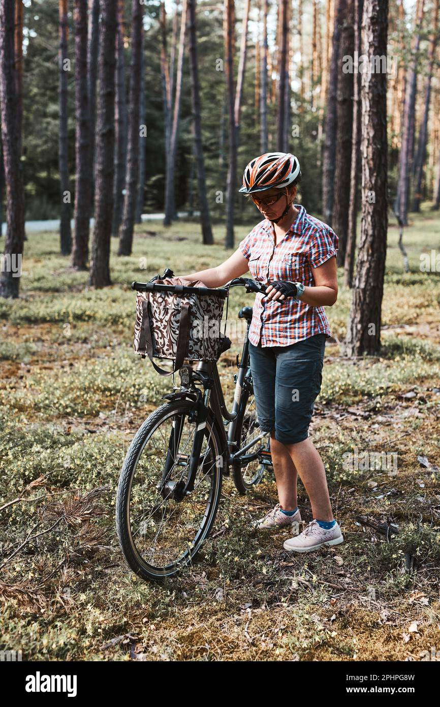 Femme active qui passe des vacances d'été gratuites lors d'un voyage à vélo dans une forêt. Femme portant un casque de vélo et des gants tenant un vélo avec un panier Banque D'Images