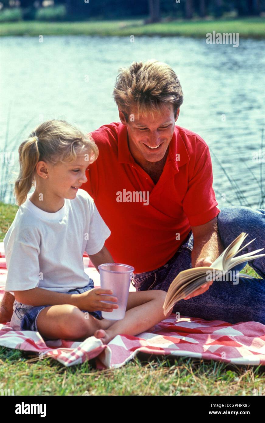 1990 PÈRE HISTORIQUE ENFANT ASSIS PRÈS DU LAC À L'EXTÉRIEUR DE L'ENSEIGNEMENT DE LA LECTURE ENSEMBLE PAR LE LAC Banque D'Images