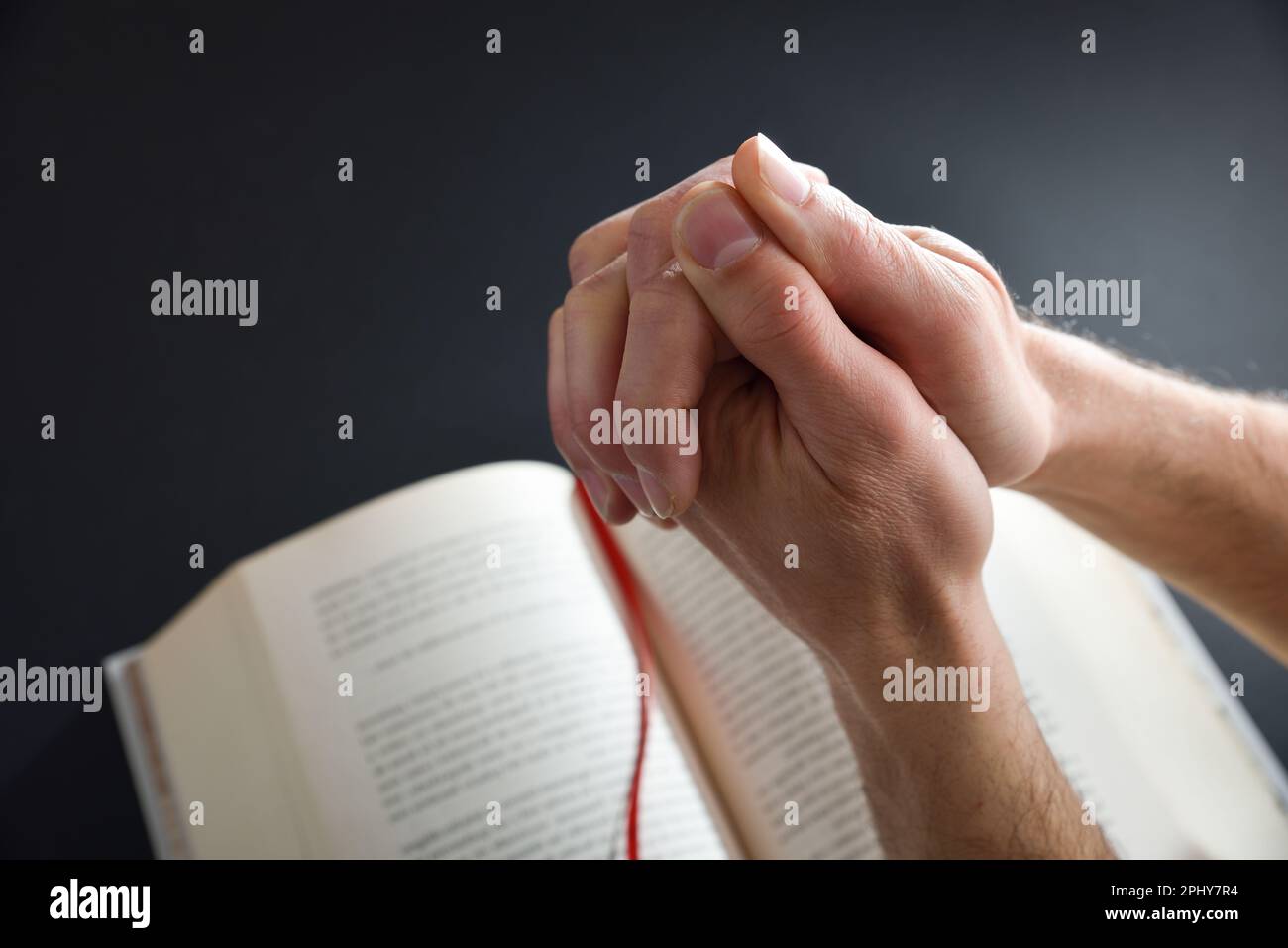 Homme religieux priant avec les mains et les doigts entremêtés et livre religieux en arrière-plan sur table noire. Vue de dessus. Banque D'Images