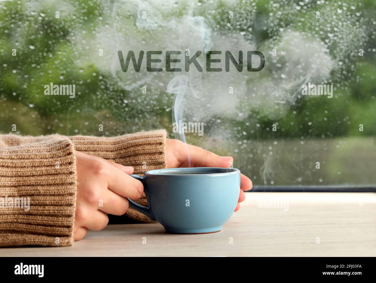 Bonne fin de semaine. Femme avec une tasse de boisson chaude à une table en bois près de la fenêtre le jour de la pluie, gros plan Banque D'Images