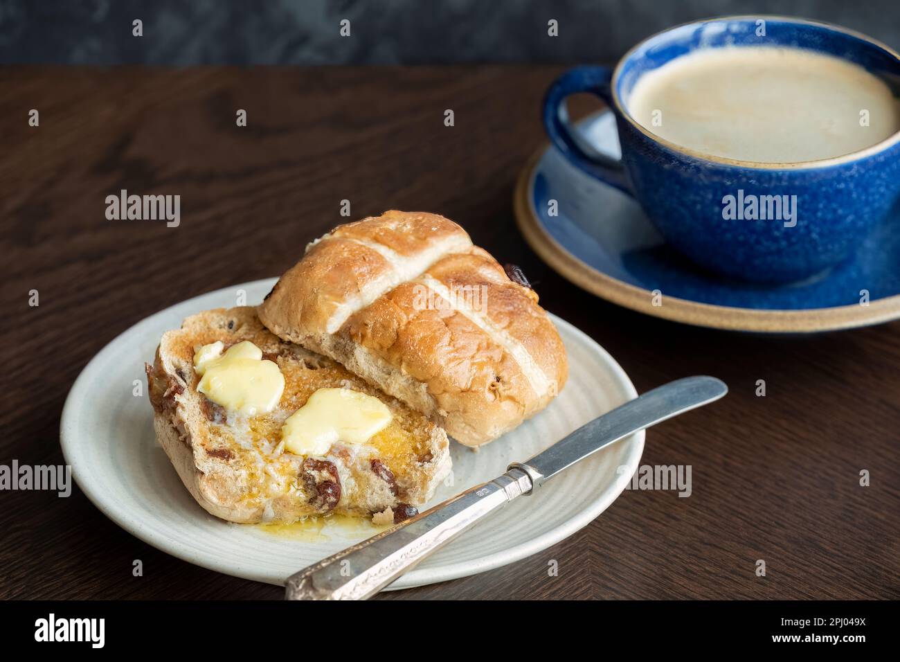 Un pain chaud croisé coupé en deux et étalé avec du beurre qui fond dans le pain. Le gâteau est servi avec une tasse de café. Un festin du Vendredi Saint Banque D'Images