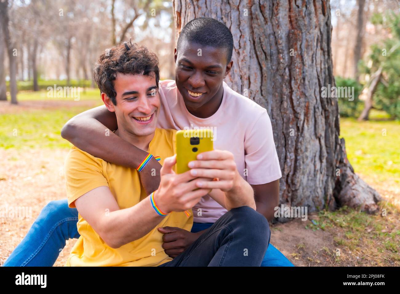 Couple d'hommes multiethniques dans un parc, concept lgbt, assis à côté d'un arbre ayant le plaisir avec un mobile jaune Banque D'Images