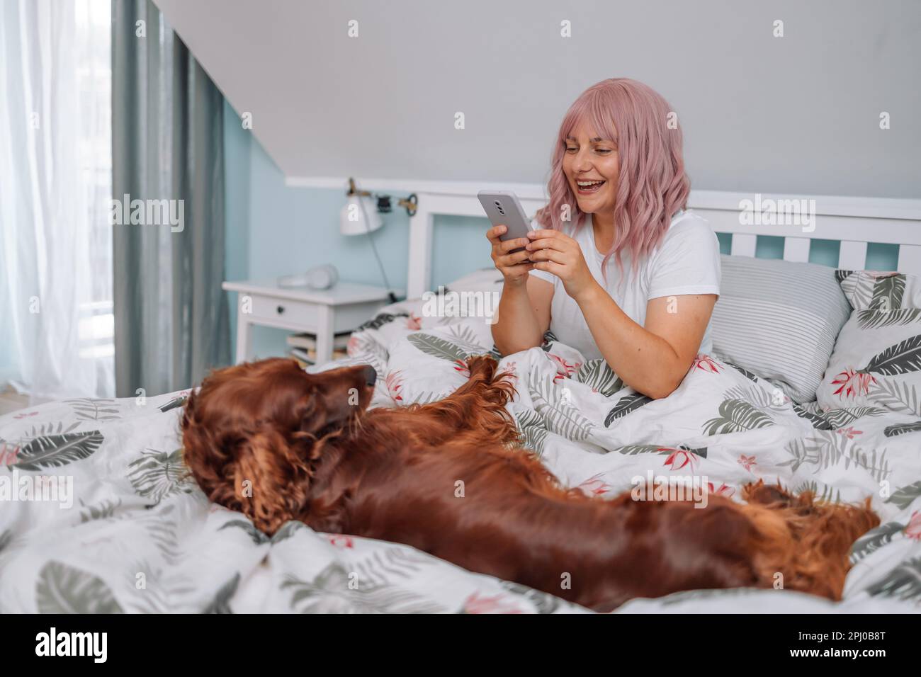 Une jeune femme prend une photo par téléphone et son chien Setter irlandais  au lit à la maison. Portrait d'une jeune femme à cheveux roses en pyjama  rose pâle avec un chien