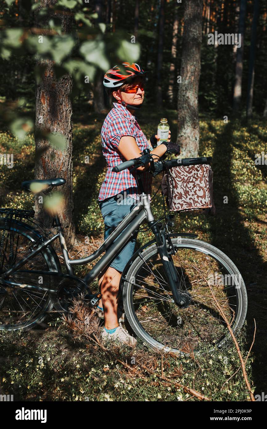 Femme active qui passe des vacances d'été gratuites lors d'un voyage à vélo dans une forêt. Femme portant un casque de vélo et des gants tenant un vélo avec un panier et un dr Banque D'Images